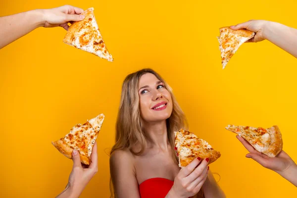 A beautiful sensual fit young blonde girl surrounded with hands with pizza slices around her. Isolated on yellow.