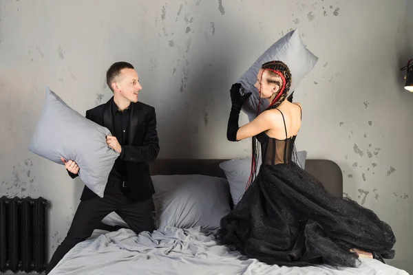 A graceful and elegant woman in stylish black dress with long red braids and man in suit are fighting with pillows.