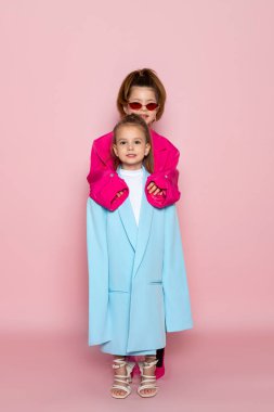 Yetişkinler gibi giyinmiş iki küçük kız. Büyük beden kıyafetler giyen çocuklar. Modaya uygun çocuklar. Pembe arka planda izole edilmiş.
