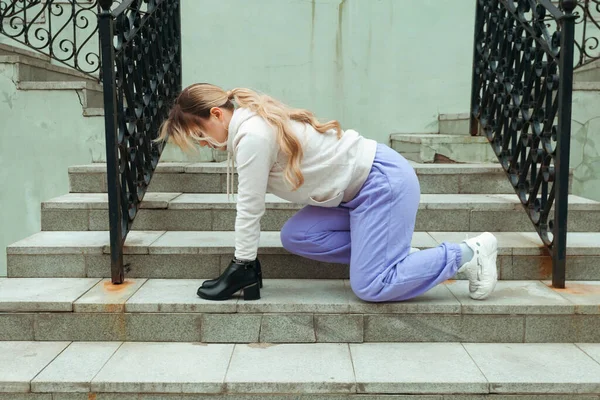 Street photography. A young blonde girl in a street sportswear is sitting on building stairs and wearing a shoes on her hands.