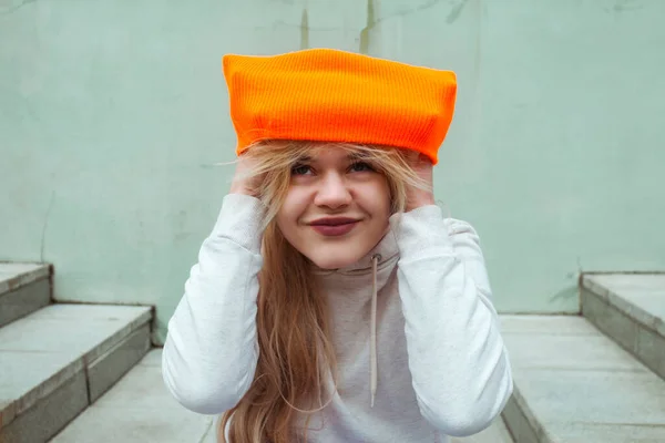 Street photography. A young blonde girl in a street sportswear is sitting on building stairs and putting on a bright orange beanie or hat.