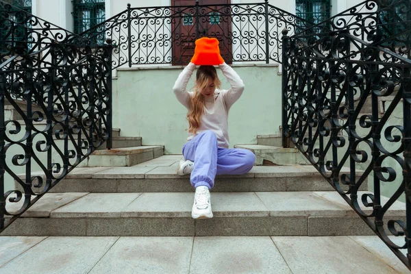 Street photography. A young blonde girl in a street sportswear is sitting on building stairs and putting on a bright orange beanie or hat.
