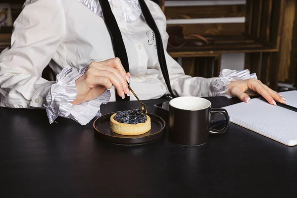 A beautiful woman in a dressy vintage shirt sitting by the black wooden desk, working, and eating a small blueberry cake.