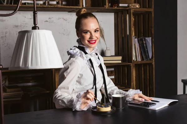 A beautiful woman in a dressy vintage shirt and bright red lipstick sitting by the black wooden desk, working, and eating a small blueberry cake.