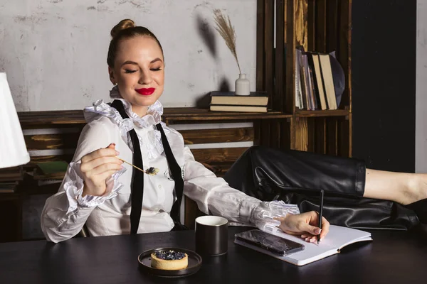 A beautiful woman in a dressy vintage shirt and bright red lipstick sitting by the black wooden desk, working, and eating a small blueberry cake.