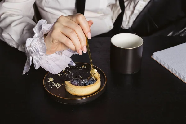 A beautiful woman in a dressy vintage shirt sitting by the black wooden desk, working, and eating a small blueberry cake.