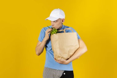 Mavi tişörtlü ve şapkalı genç adam içinde yiyecek olan bir kese kağıdı tutuyor. Sarı arka planda izole edilmiş. Teslimat konsepti.