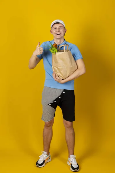 青いTシャツとキャップを着た若い男が食べ物の入った紙袋を持っています 黄色の背景に隔離されている デリバリーコンセプト — ストック写真