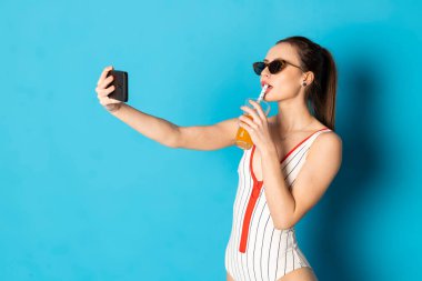 Şehvetli beyaz bir mayo giymiş güzel, mutlu genç bir kadın ve pipetten limonata içerken selfie çekiyor. Mavi arkaplanda izole.