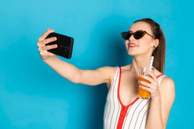 Şehvetli beyaz bir mayo giymiş güzel, mutlu genç bir kadın ve pipetten limonata içerken selfie çekiyor. Mavi arkaplanda izole.