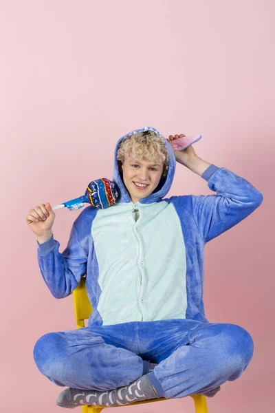 一个卷曲的金发小伙子穿着可爱可笑的蓝色睡衣假装是个孩子 手里拿着一个大棒棒糖 以粉红为背景隔离 — 图库照片