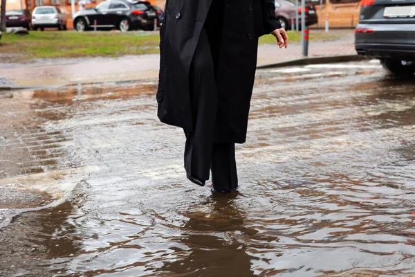 An well dressed young woman in black coat and cannot cross the road because of a large puddle. The flooded street does not allow pedestrians to pass.