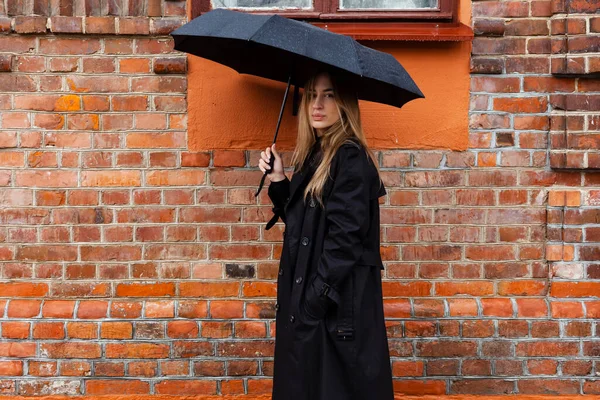 黒の美しい少女の肖像画 コートに身を包んだと傘を保持 雨の日に明るいオレンジ色のレンガの壁に対して通りを下って行く — ストック写真