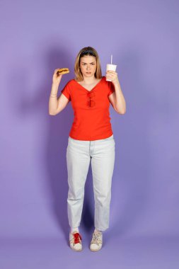 Mutsuz, üzgün, güzel bir kız fast food yiyor, ucuz burger yiyor, elinde çizburger ve soda tutuyor. Fast food ve sağlıksız diyet. mor arkaplanda izole.