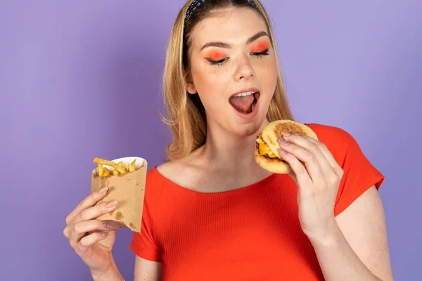 兴奋的漂亮女孩吃快餐 便宜的汉堡包 拿着薯条和汉堡包 张开嘴 快餐和不健康的饮食 紫色背景的孤立物 — 图库照片