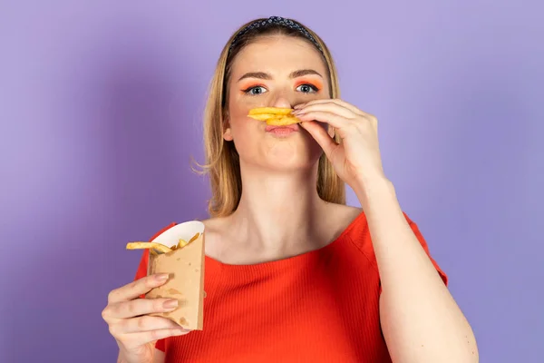 饥饿的漂亮姑娘手里拿着快餐 油腻地吃着土豆 用薯条模仿小胡子 快餐和不健康的饮食 紫色背景的孤立物 — 图库照片