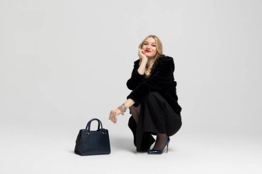 Genç kıvırcık sarışın kız şık giyinmiş, oturuyor ve bir el çantasıyla poz veriyor. Pahalı kıyafetler, tarz ve moda konsepti.