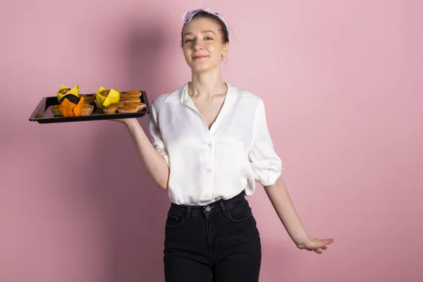 Stock image Cute happy girl is holding a tray with fresh, delicious muffins and pastries. Isolated on pink background.