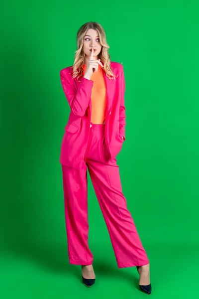 stock image Young curly blonde dressed in pink blazer and pants, gesturing quiet sign with a hand. Isolated on green background.
