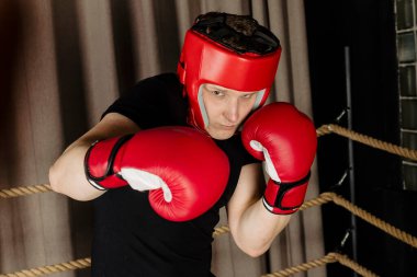 Kırmızı başlık ve eldiven takan, ringde poz veren, kameraya bakıp yumruk atan sert bir adamın portresi.