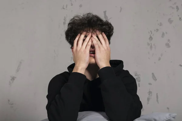 A severely depressed teenager, sitting on the bed, crying and covering his eyes. A young man in a hard mental condition.