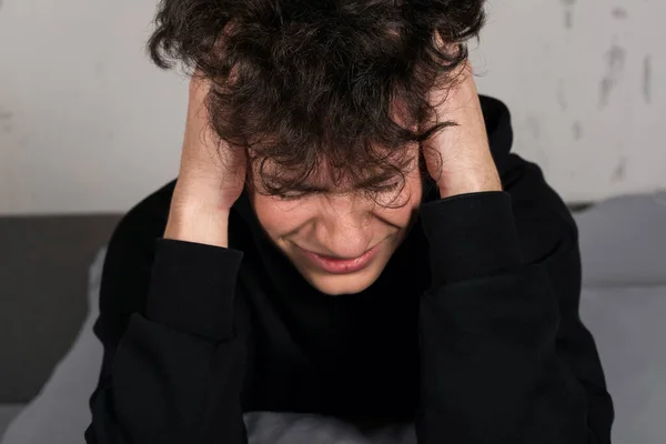 A severely depressed teenager, sitting on the bed, holding his head. A young man in a hard mental condition.