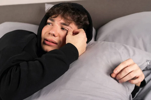 A severely depressed teenager, lies on the bed, embracing a pillow and crying with teary eyes. A young man in a hard mental condition.