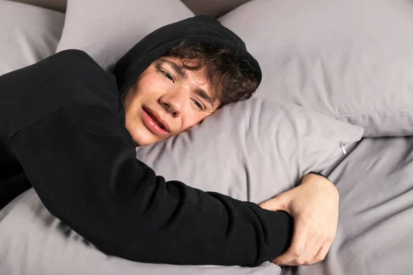 A severely depressed teenager, lies on the bed, embracing a pillow and crying with teary eyes. A young man in a hard mental condition.