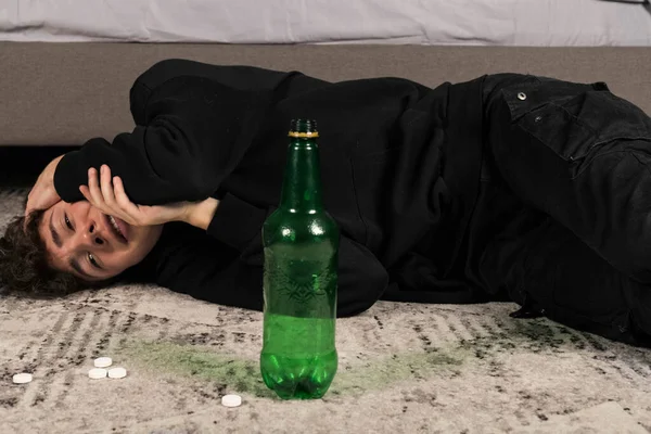 A severely depressed guy, lying on the floor among pills and a bottle of alcohol. A young man in a hard mental condition. alcohol and drugs addiction.