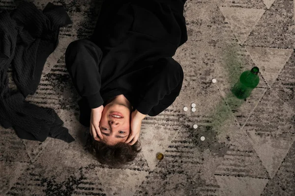 A severely depressed guy, lying on the floor among pills and a bottle of alcohol. A young man in a hard mental condition. alcohol and drugs addiction. Top view. view from above.