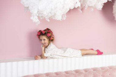 Saç lastiği takan, beyaz benekli elbise giyen, bulutların altında yatan küçük bir kız. Pastel pembe arkaplan.