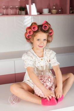 Saç bigudisi ve beyaz benekli elbise giyen küçük bir kız, pembe bir mutfak masasında oturur, büyük cam bir kavanozla şekerleme ya da şekerlemelerle.