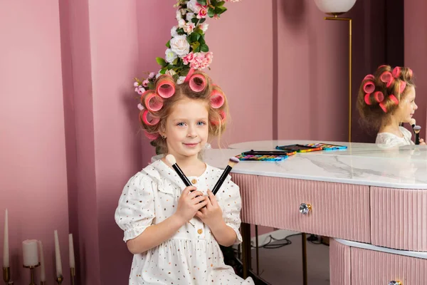 Little girl wearing hair rollers and white polka dot dress sits in the pastel pink dressing room by a mirror, holds cosmetics and a makeup brush