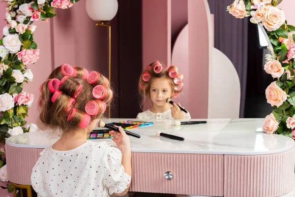 Little girl wearing hair rollers and white polka dot dress sits in the pastel pink dressing room by a mirror, holds cosmetics and a makeup brush