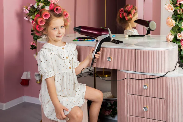 Little girl wearing hair rollers and white polka dot dress sits in the pastel pink dressing room by a mirror, holds a hair dryer.