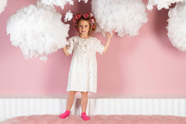 Little girl wearing hair rollers and white polka dot dress reaching her hands up to the clouds. Child touches cloud. pastel pink colored background.