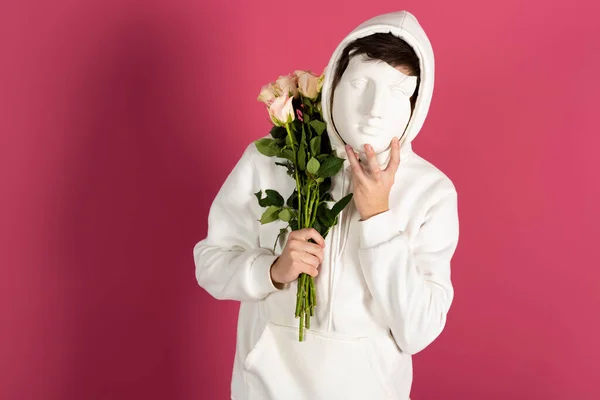 stock image Suspicious young man in white hoodie, holds bouquet of flowers or roses and covering face with a mask. Secret admirer, Hidden identity concept. Isolated on pink background.