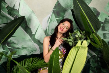 Çiçeklerin tadını çıkaran taze yeşil makyajlı güzel esmer kadın büyük tropikal yapraklarla bitkilerin arasında poz veriyor. Doğa kavramına saygı göstermek..