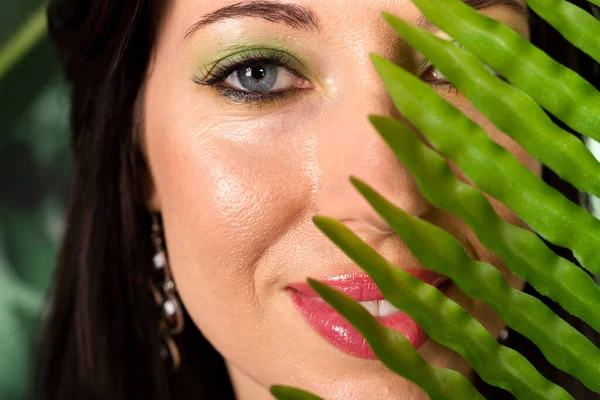 Half covered face close-up. Beautiful brunette woman with fresh green makeup enjoying the floral environment, posing among plants with big tropical leaves. Respecting the nature concept.