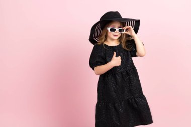 Siyah şık bir yaz elbisesi, güneş şapkası ve havalı beyaz güneş gözlüğü takan sevimli küçük bir kız. Pembe arka planda izole edilmiş.