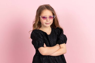 Siyah şık bir yaz elbisesi, makyajı ve pembe ince güneş gözlüğü olan sevimli küçük bir kız. Pembe arka planda izole edilmiş.