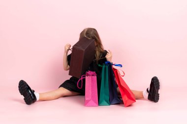 Çocuk alışverişi. Siyah şık bir yaz elbisesi ve hafif makyajlı sevimli küçük bir kız yerde oturmuş alışveriş poşetlerine bakıyor. Pembe arka planda izole edilmiş.