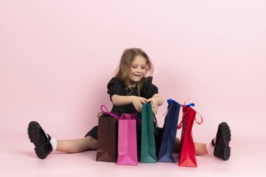 Çocuk alışverişi. Siyah şık bir yaz elbisesi ve hafif makyajlı sevimli küçük bir kız yerde oturmuş alışveriş poşetlerine bakıyor. Pembe arka planda izole edilmiş.