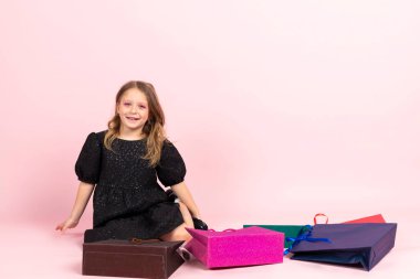 Çocuk alışverişi. Siyah şık bir yaz elbisesi ve hafif makyajlı sevimli küçük bir kız yerde oturmuş alışveriş poşetlerine bakıyor. Pembe arka planda izole edilmiş.