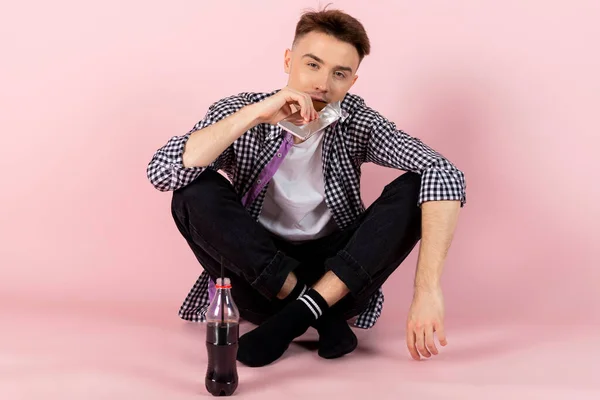 A happy young man drinking coke and eating a chocolate bar. fast food and sugar addiction, unhealthy diet. Isolated on pink background.