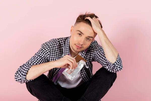 A happy young man drinking coke and eating a chocolate bar. fast food and sugar addiction, unhealthy diet. Isolated on pink background.