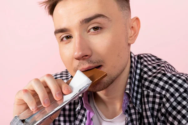 A happy young man drinking coke and eating a chocolate bar. fast food and sugar addiction, unhealthy diet. Isolated on pink background.