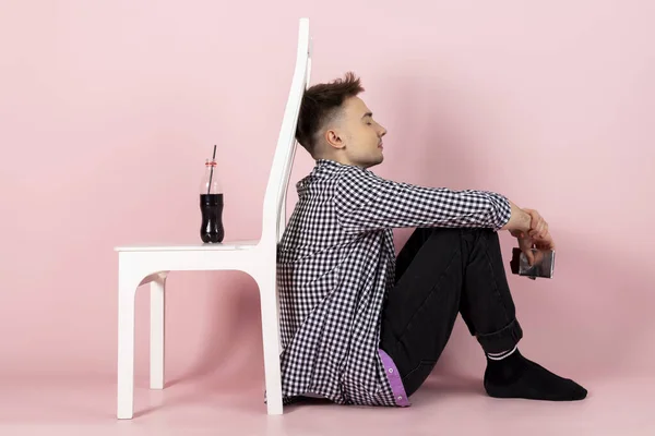 A young man holding a chocolate bar and turned his back to a bottle of coke or sweet soda. fast food and sugar addiction, unhealthy diet. Isolated on pink background.
