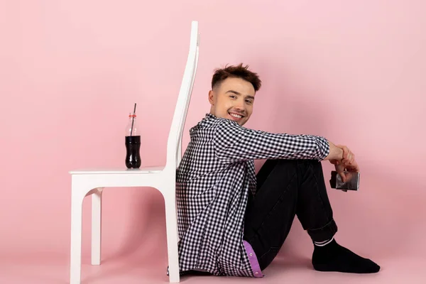 A young man holding a chocolate bar and turned his back to a bottle of coke or sweet soda. fast food and sugar addiction, unhealthy diet. Isolated on pink background.
