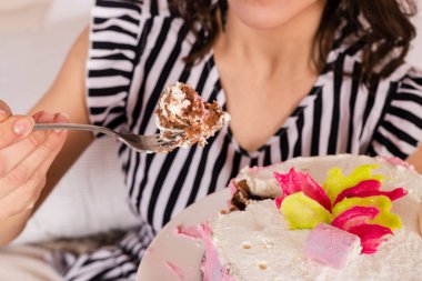 Güzel esmer kadın doğum gününü kutluyor, çizgili kısa bir elbise ve doğum günü şapkası giyiyor, yatağa oturuyor ve pembe pastadan bir parça yiyor.